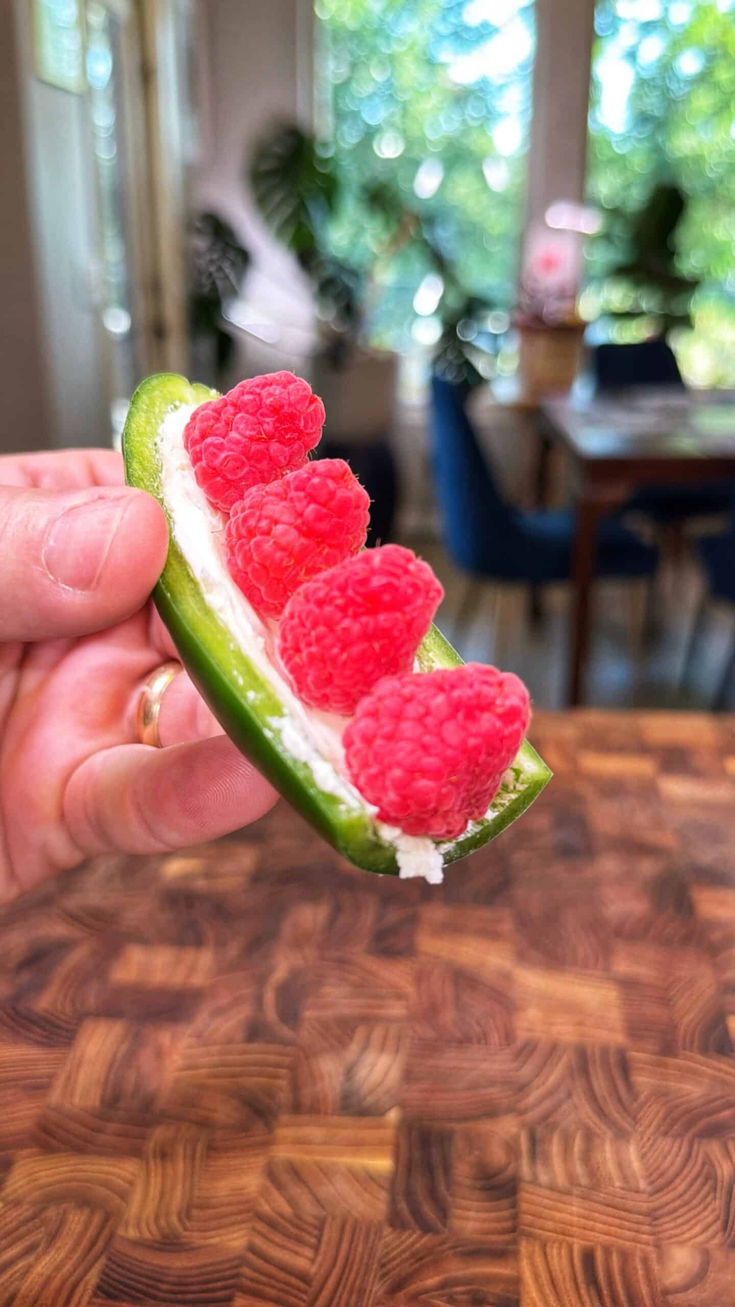 A hand holds a halved jalapeño filled with cream cheese and topped with four fresh raspberries; a kitchen with plants and large windows is blurred in the background.