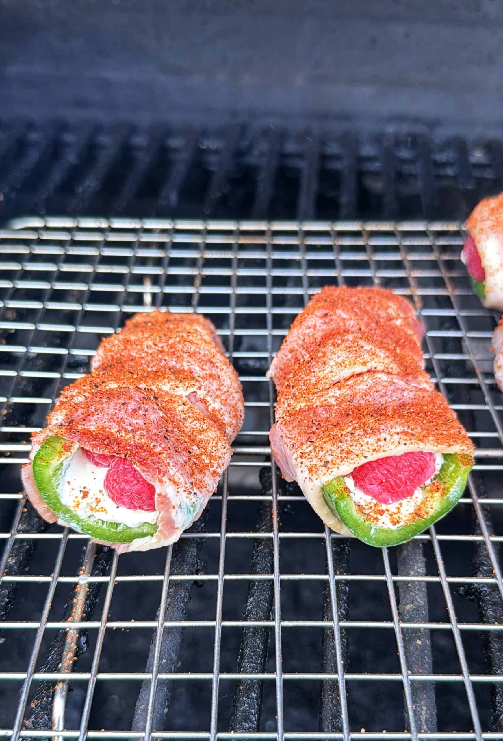 Two bacon-wrapped jalapeño poppers filled with cream cheese and a raspberry, seasoned with spices, are grilling on a wire rack.