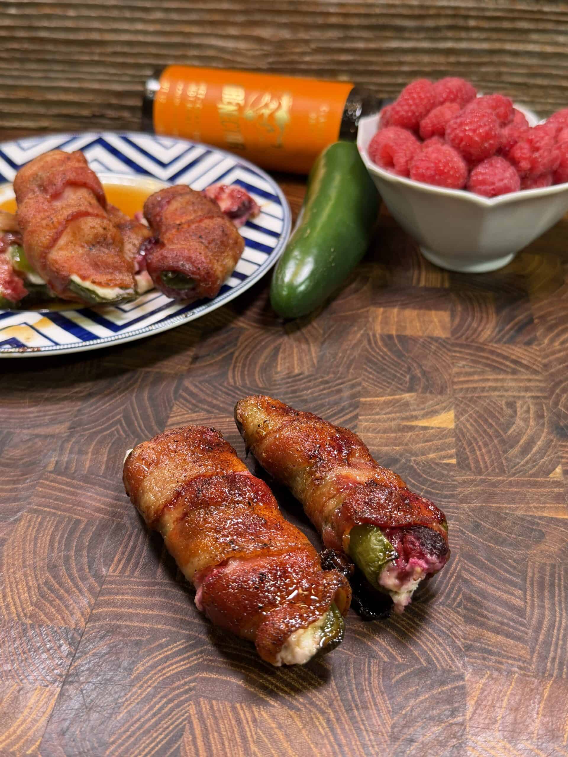 Two bacon-wrapped jalapeño poppers rest on a wooden board, with a bowl of raspberries, whole jalapeño, a plate of more poppers, and an orange sauce bottle in the background.
