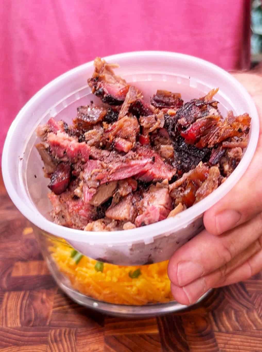 A hand holds a plastic container filled with chopped smoked brisket above another bowl containing shredded cheese and green onions.