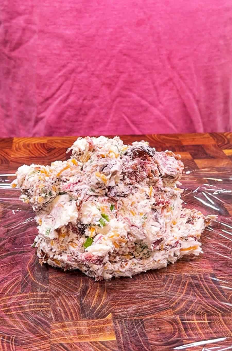 A mound of mixed cheese, herbs, and meat chunks sits on a wooden surface with plastic wrap, against a pink fabric background.