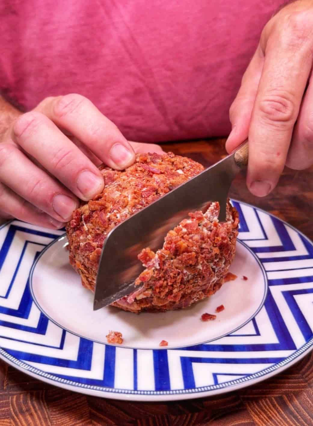 A person cuts into a round, crispy, bacon-covered food item on a decorative plate with a large knife.