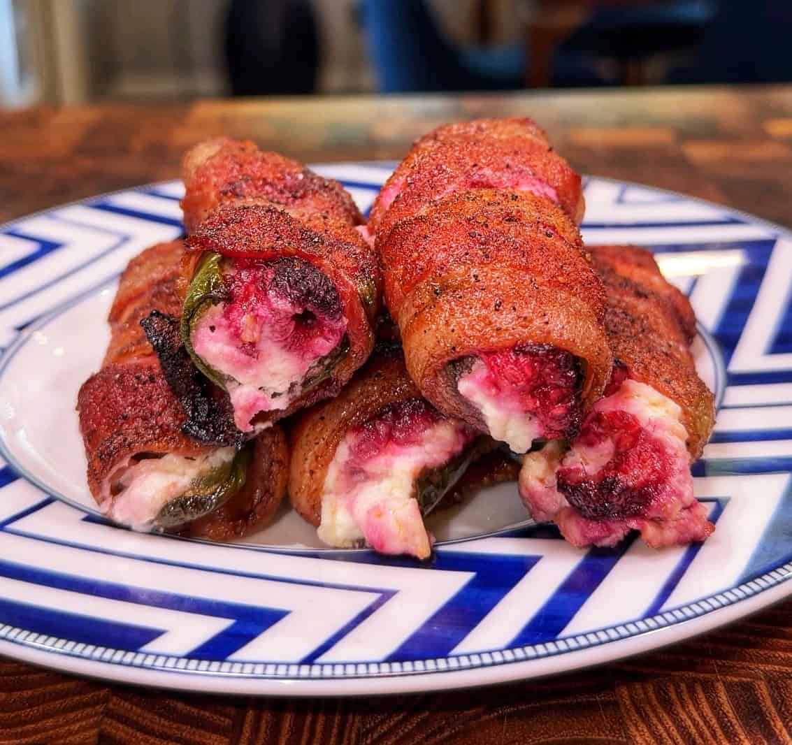 A plate of bacon-wrapped jalapeños stuffed with creamy cheese and topped with a red seasoning, arranged neatly on a geometric-patterned plate.