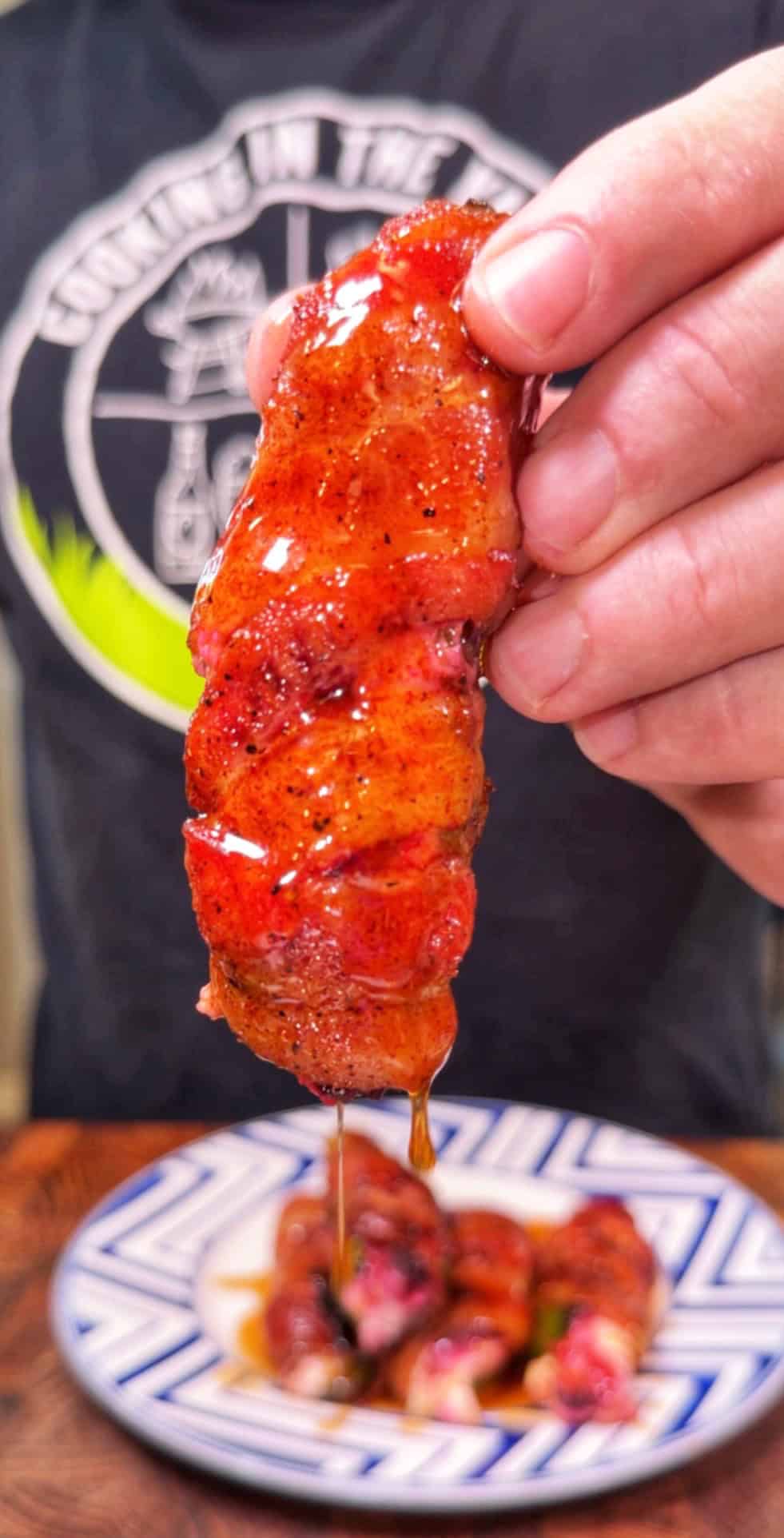 A hand holds a glazed, bacon-wrapped piece of food with sauce dripping off, over a patterned plate. A person in a dark shirt with white logo is in the background.