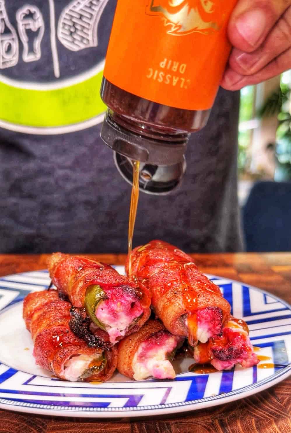 A hand pours sauce from an orange bottle onto bacon-wrapped, cheese-stuffed jalapeños stacked on a white and blue plate.