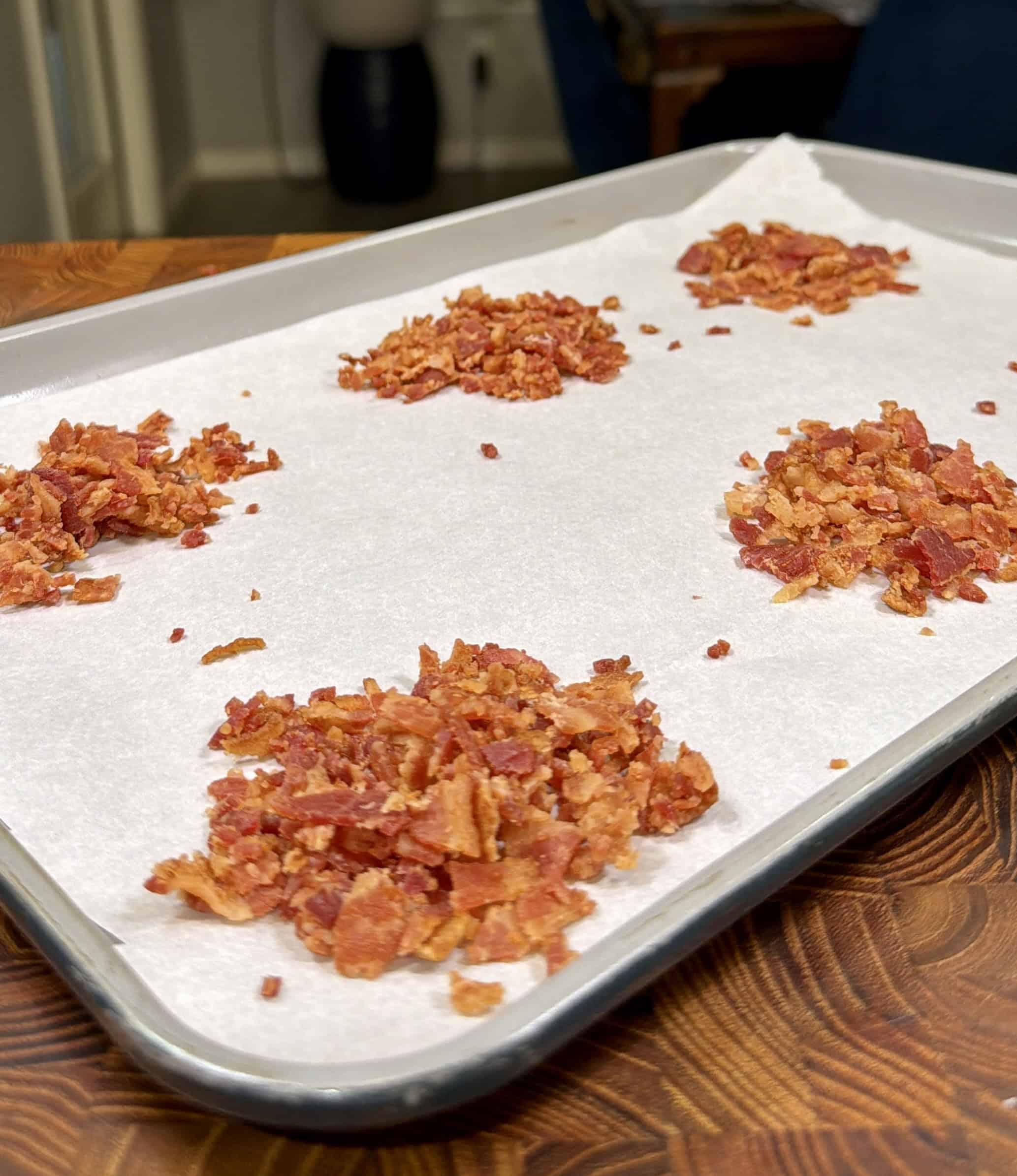 Five small piles of crumbled cooked bacon are arranged on a parchment-lined baking sheet, resting on a wooden surface in a kitchen setting.