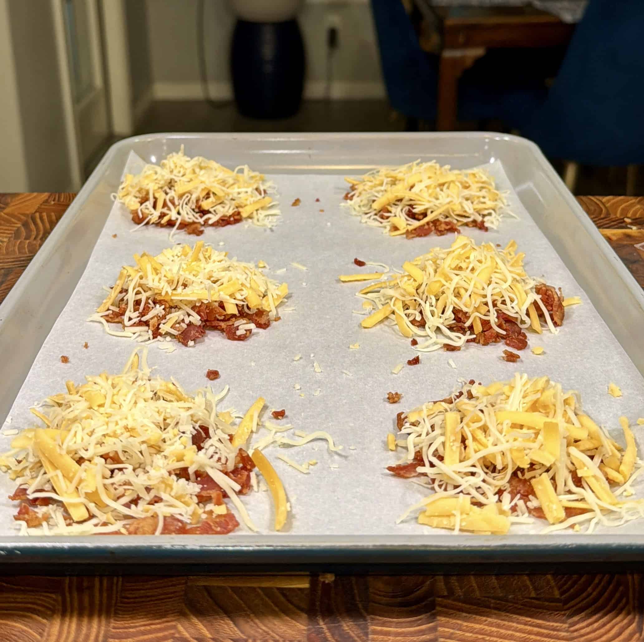 Six small piles of shredded cheese and crumbled bacon are arranged on a parchment-lined baking sheet, ready to be baked. The baking tray sits on a wooden surface in a cozy kitchen setting.