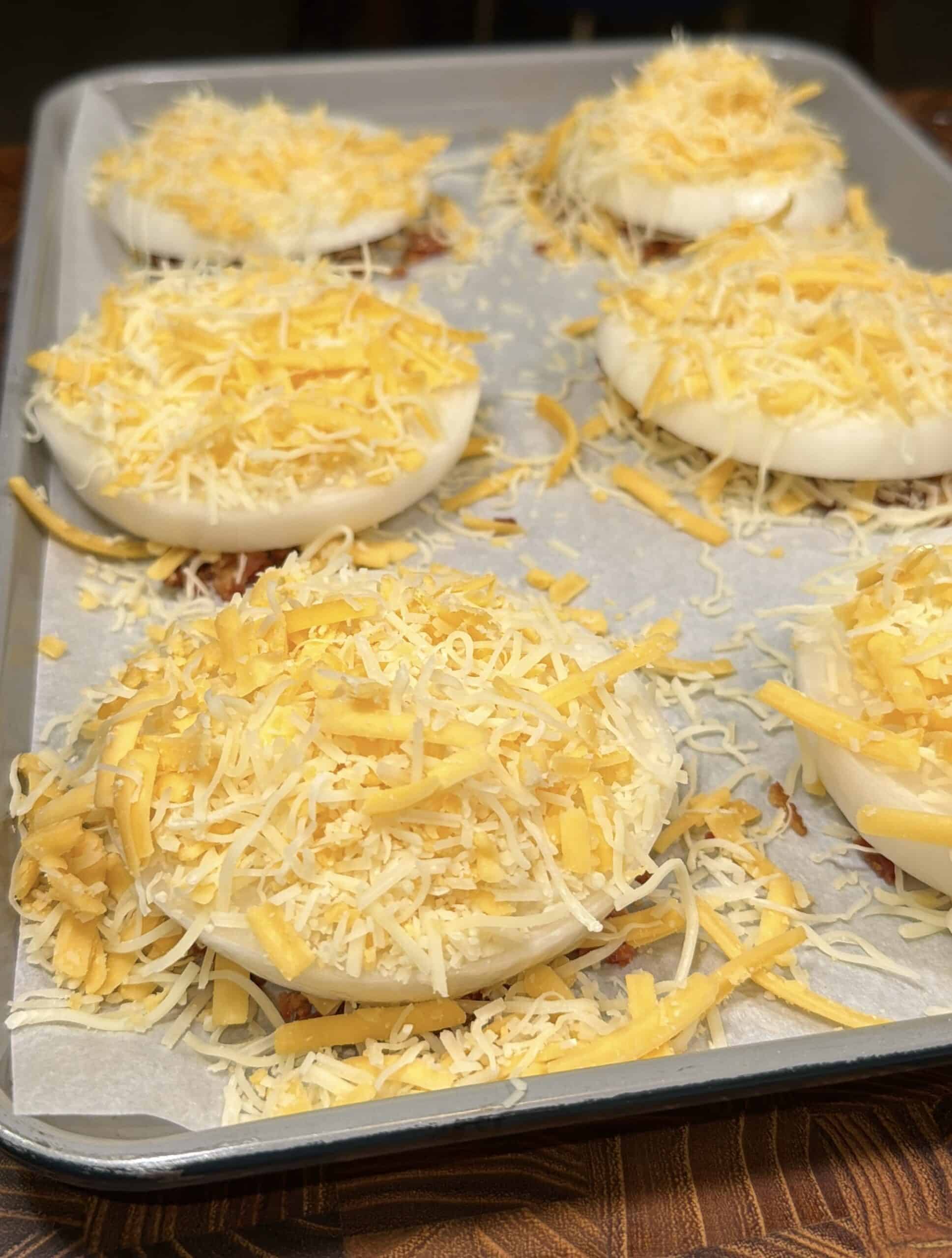 Six large onion slices topped with shredded yellow and white cheese are arranged on a parchment-lined baking sheet, ready for baking.