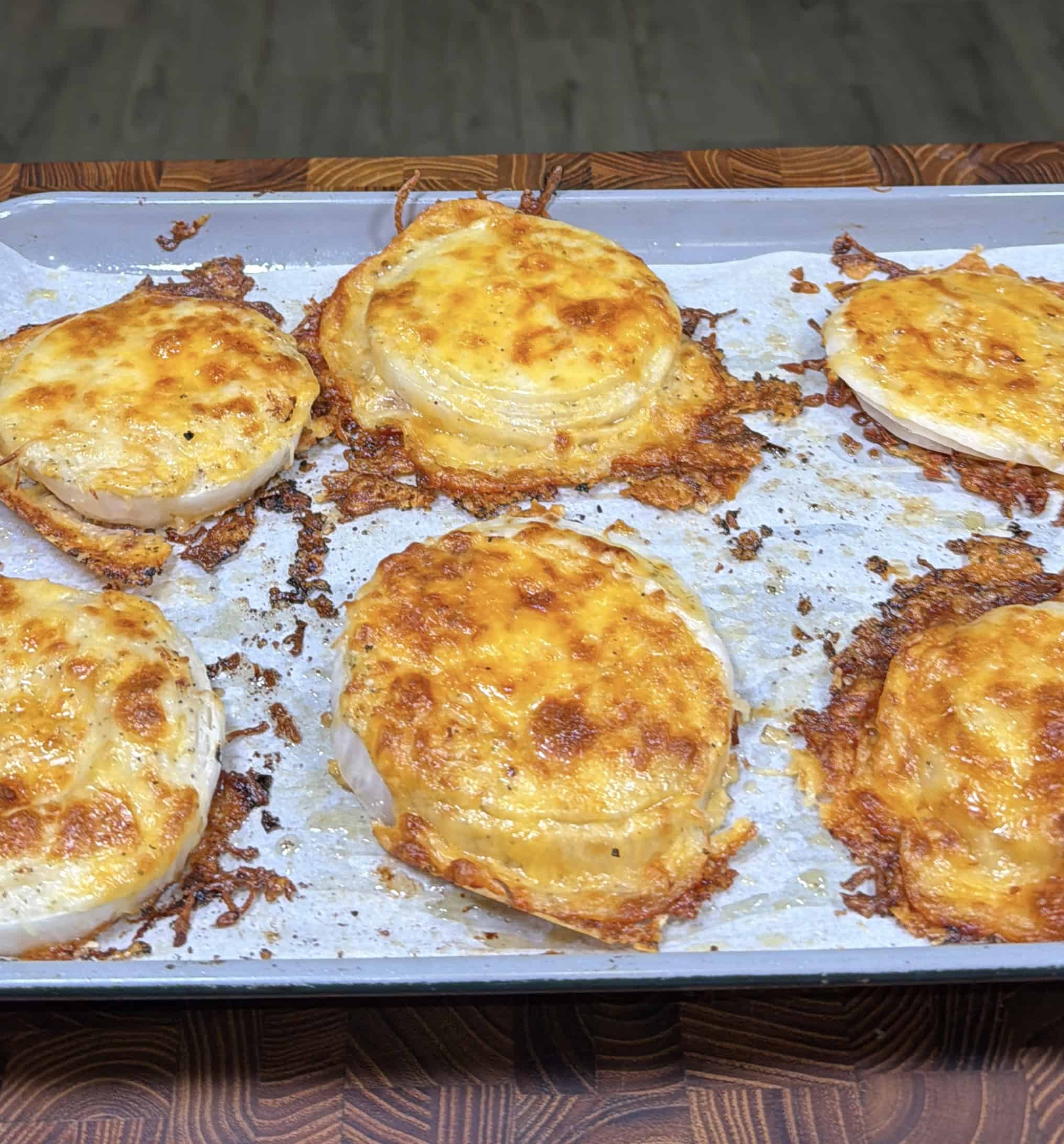 Six baked cheesy onion slices on a parchment-lined baking sheet, golden brown with melted cheese bubbling on top, resting on a wooden surface.