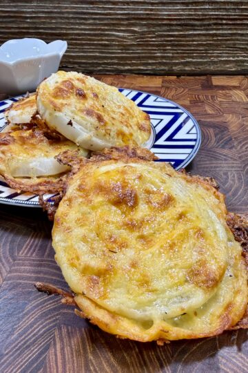 Three cheesy, golden-brown baked rounds are stacked on a patterned plate, with one in the foreground on a wooden surface. A small white bowl sits behind the plate. The background features a rustic wood texture.