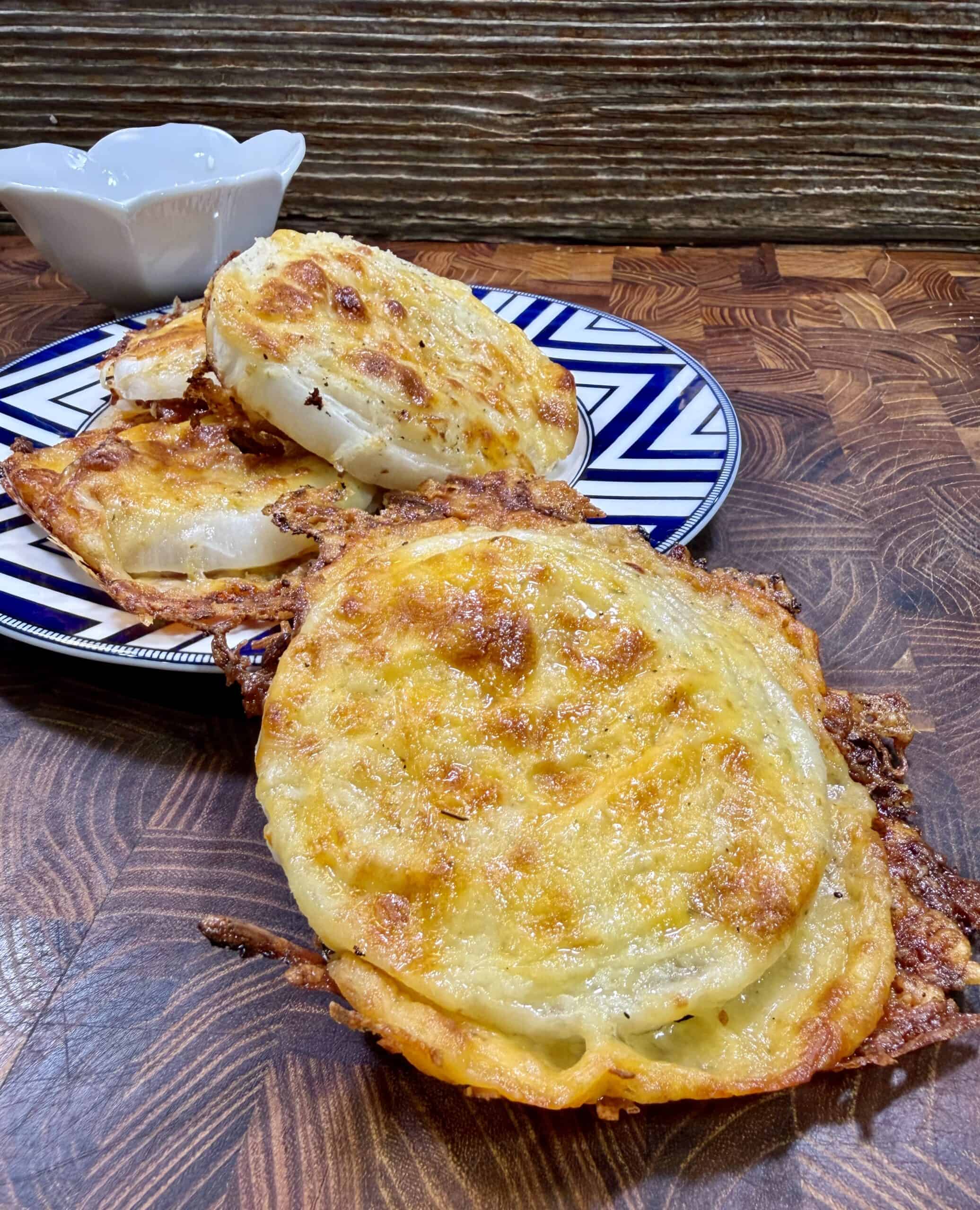 Three cheesy, golden-brown baked rounds are stacked on a patterned plate, with one in the foreground on a wooden surface. A small white bowl sits behind the plate. The background features a rustic wood texture.