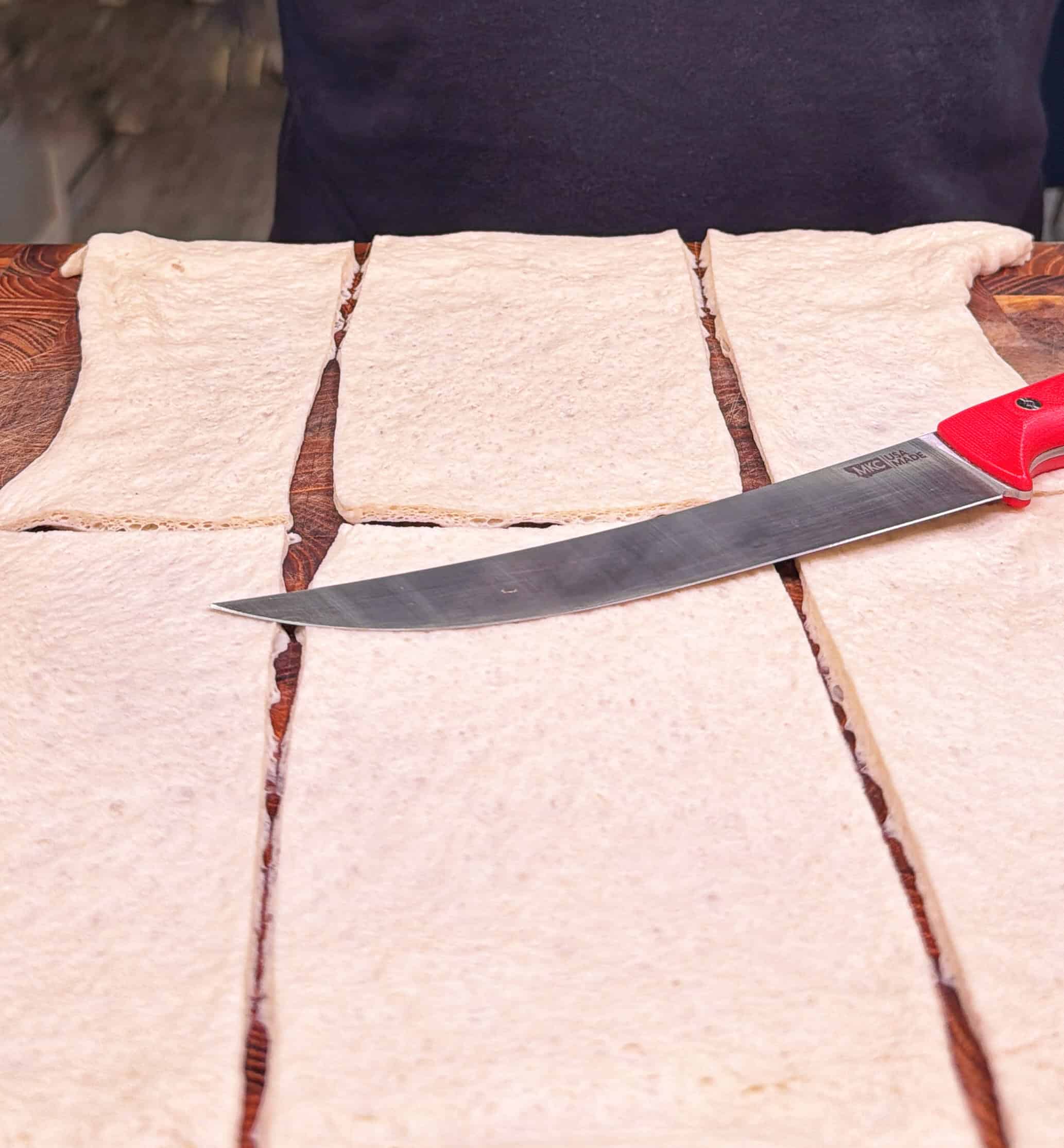 Rectangular pieces of dough are laid out on a wooden surface, with a large knife featuring a red handle resting on top. The dough appears to be evenly cut into strips.