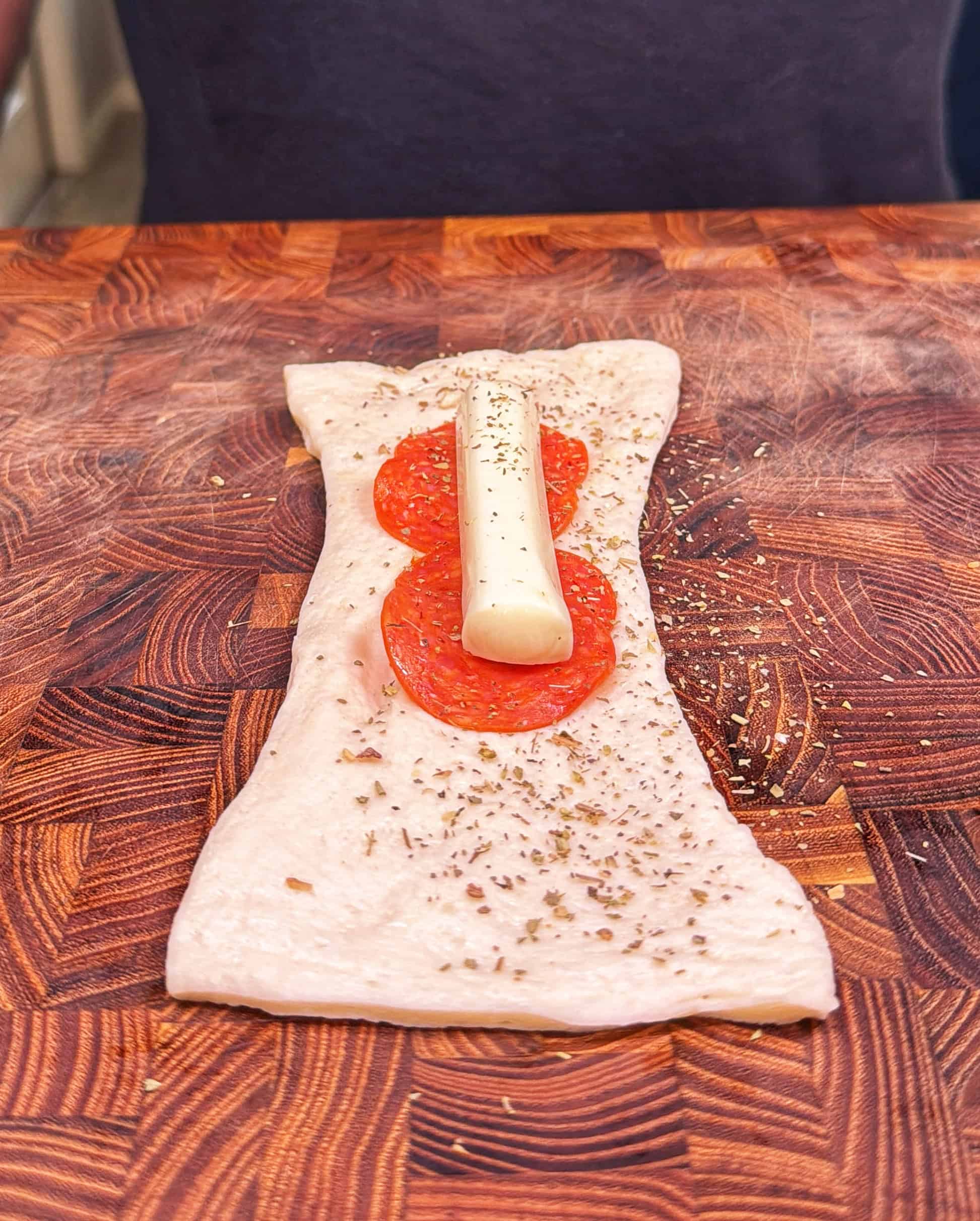 A piece of dough topped with pepperoni slices, a cheese stick, and sprinkled herbs sits on a wooden cutting board.