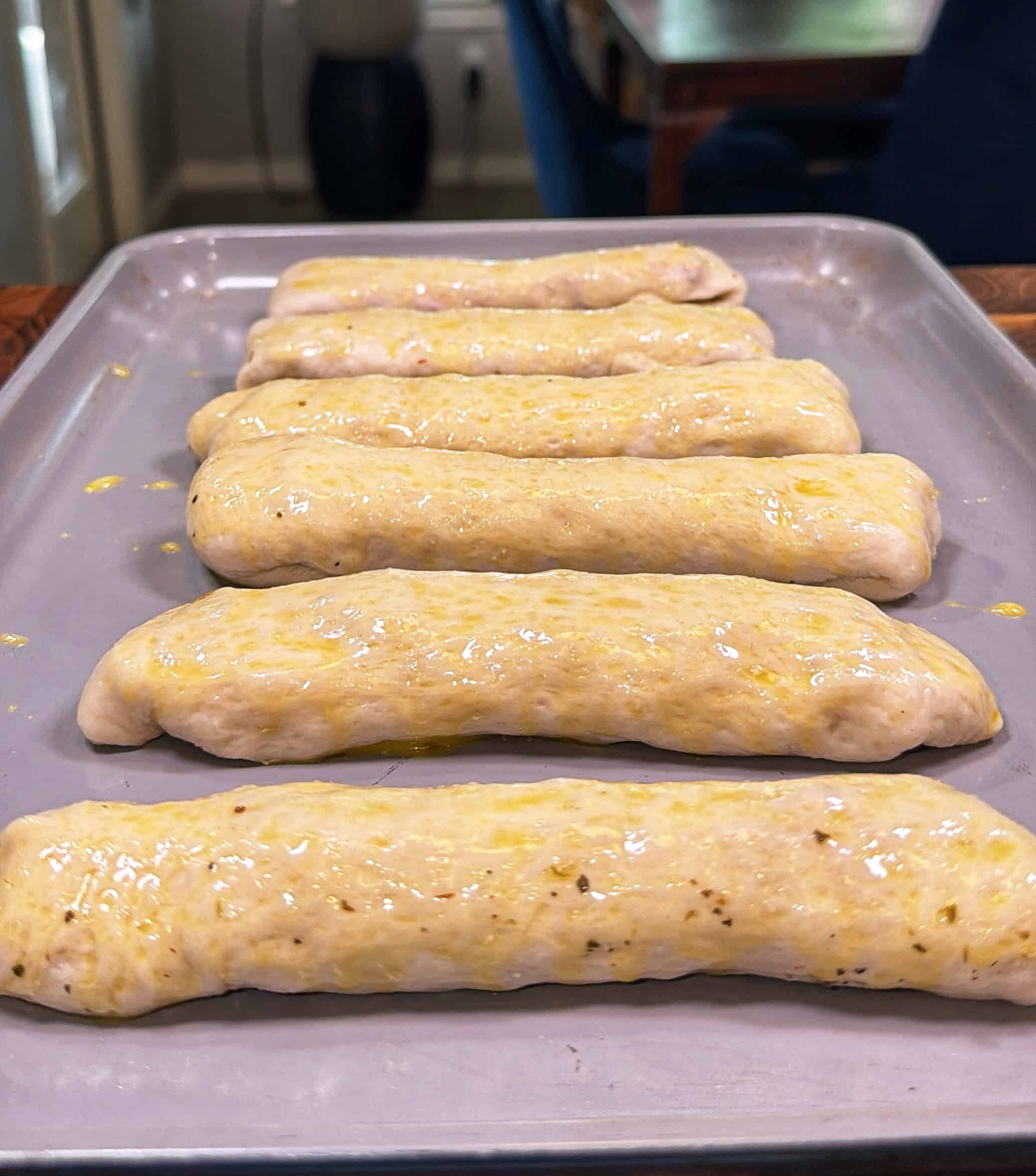Six unbaked breadsticks coated with egg wash or oil are lined up on a baking sheet, ready to be baked. The dough looks soft and slightly speckled with herbs.