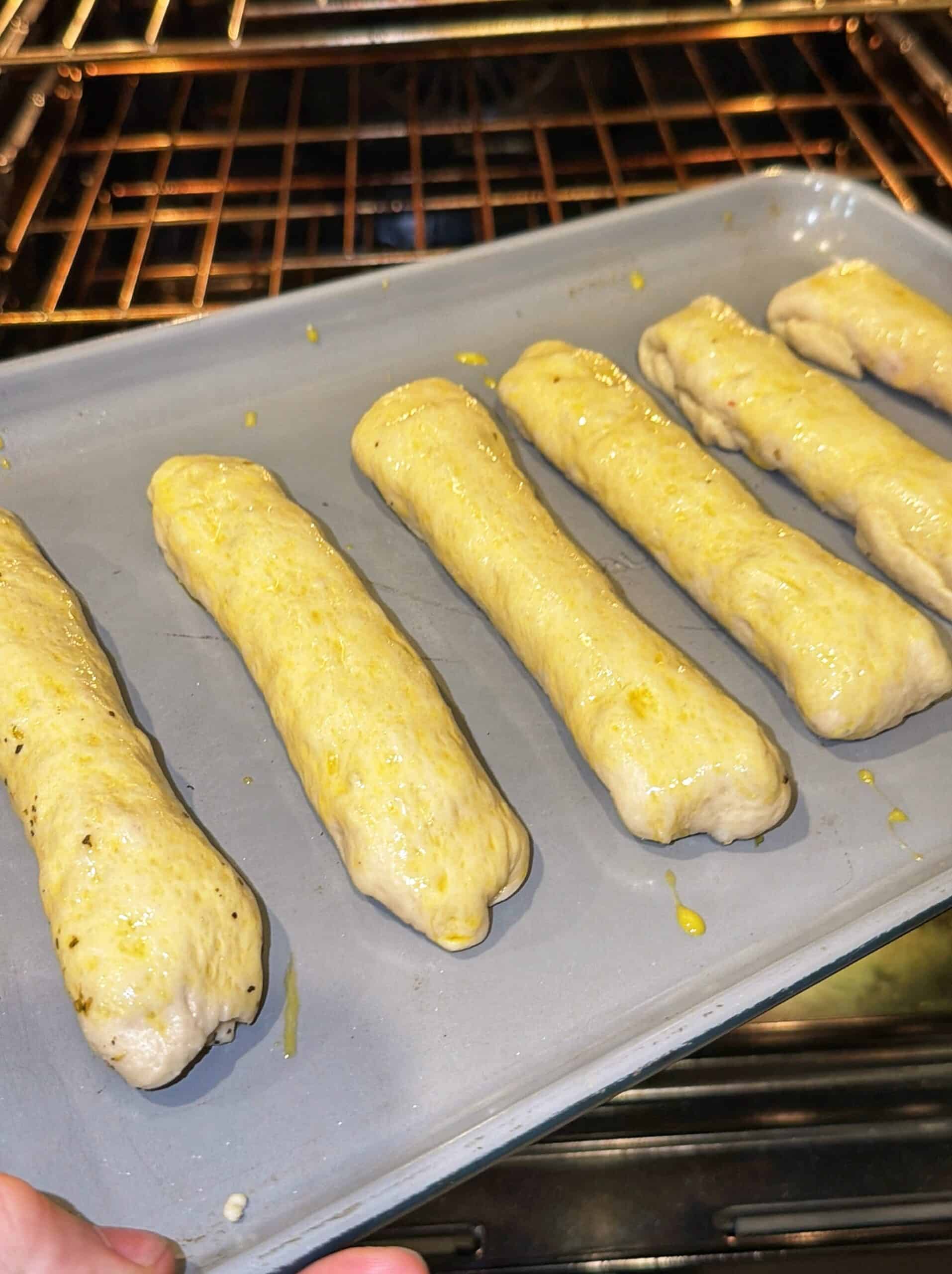 Unbaked breadsticks coated with egg wash are lined up on a baking tray, being placed into an oven with heating rods visible in the background.