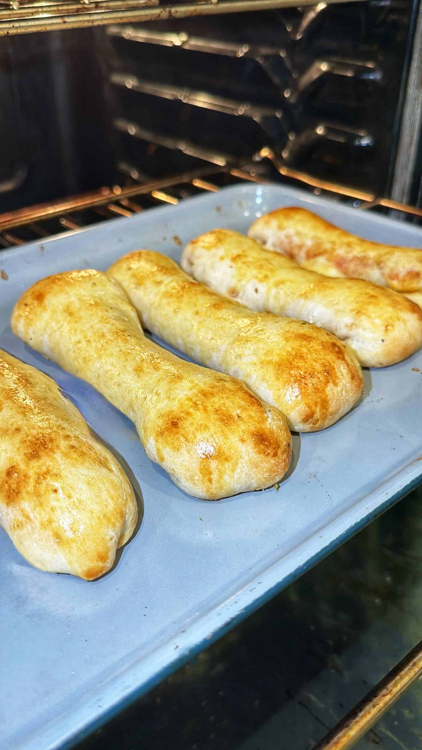 Five golden-brown breadsticks baking on a blue tray inside an oven. The breadsticks are slightly irregular in shape and have a shiny, baked finish.