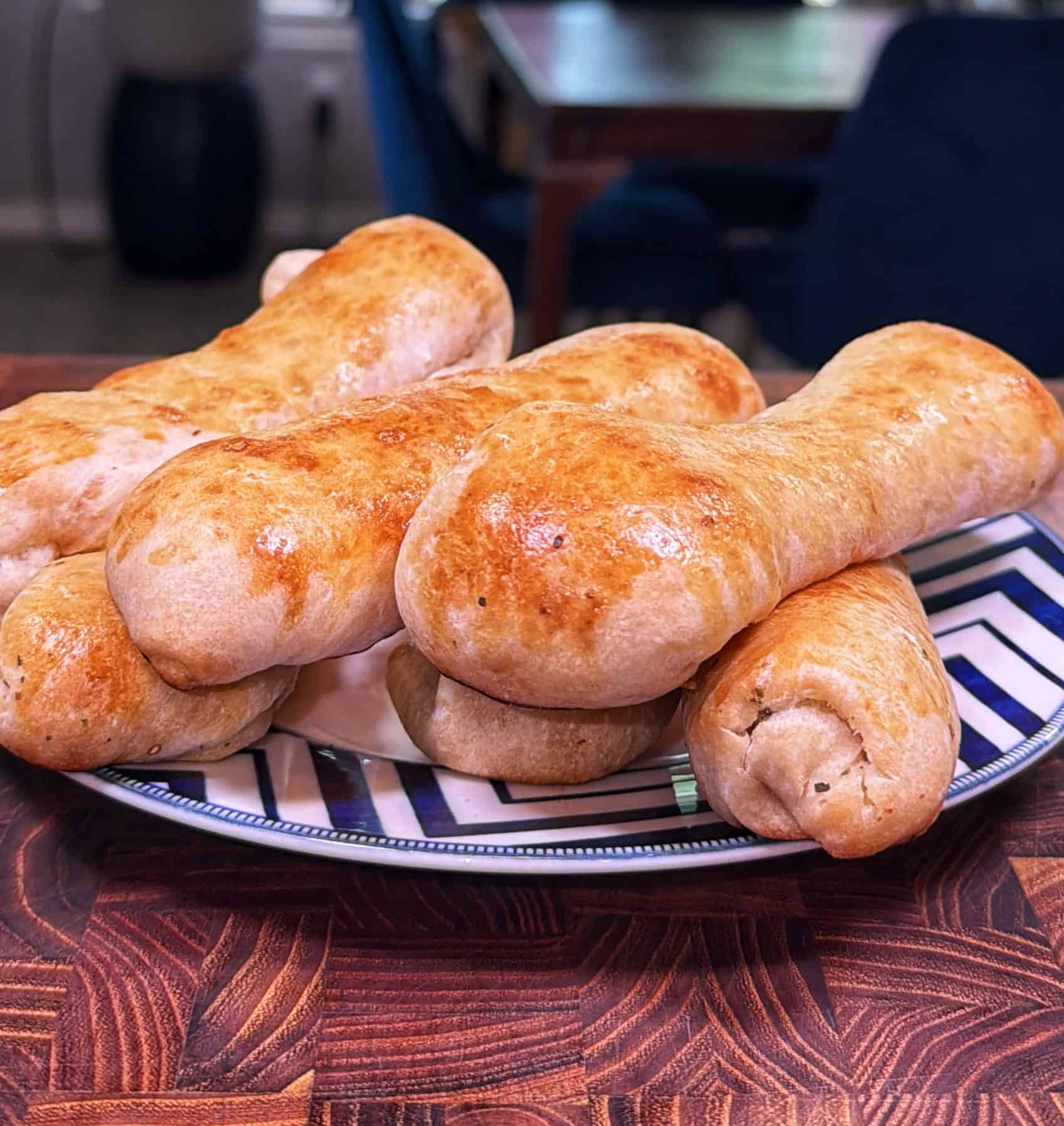 A plate of golden-brown bread rolls shaped like dog bones sits on a striped plate atop a wooden table, with a blurred dining area in the background.