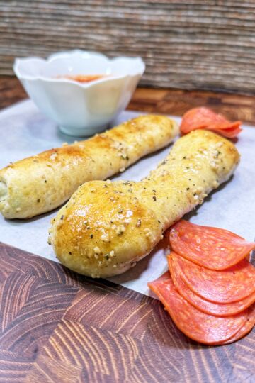 Two baked breadsticks topped with herbs and cheese, served on parchment paper. Slices of pepperoni are beside them, with a small bowl of marinara sauce in the background.