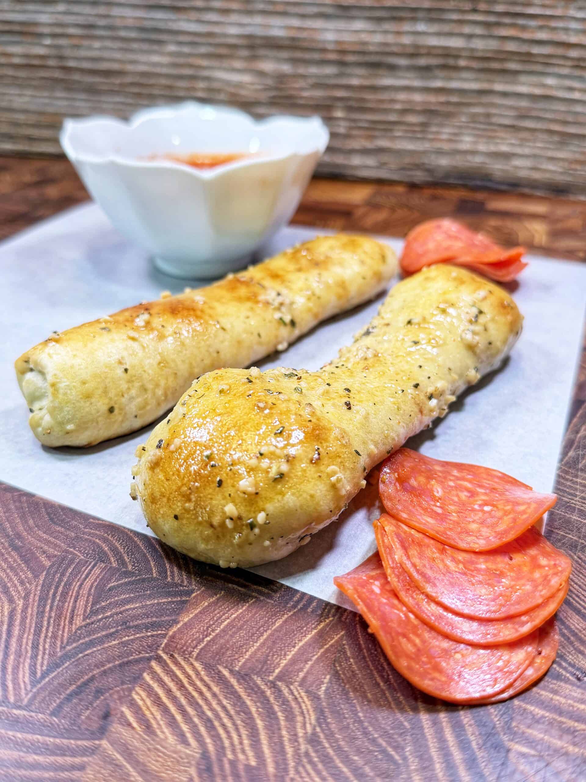 Two baked breadsticks topped with herbs and cheese, served on parchment paper. Slices of pepperoni are beside them, with a small bowl of marinara sauce in the background.