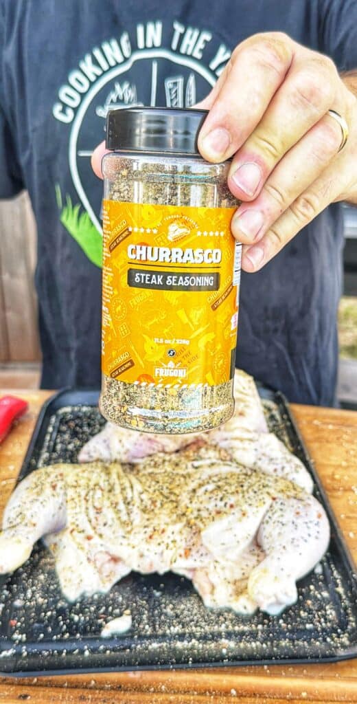 A container of seasoning held above a tray of raw seasoned frog-cut chicken.