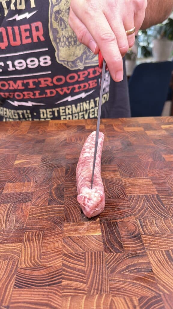 A hand slices a raw bratwurst lengthwise on a wooden cutting board.