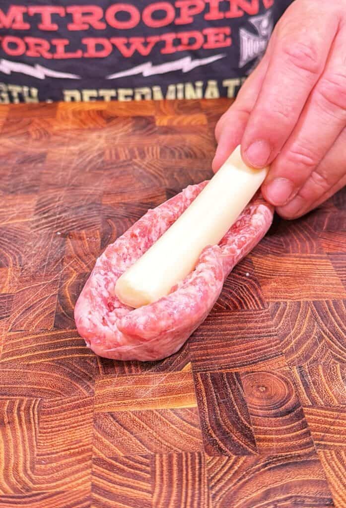 A hand places a cheese stick inside a split raw bratwurst on a wooden cutting board.