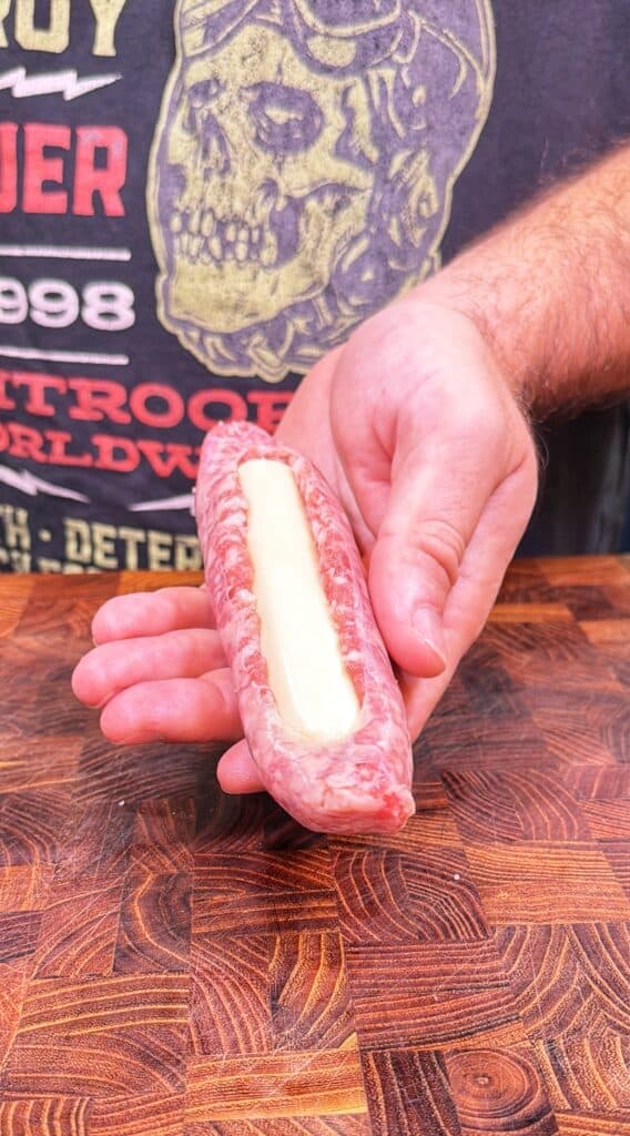 A person holds a split raw bratwurst filled with a cheese stick over a wooden cutting board.