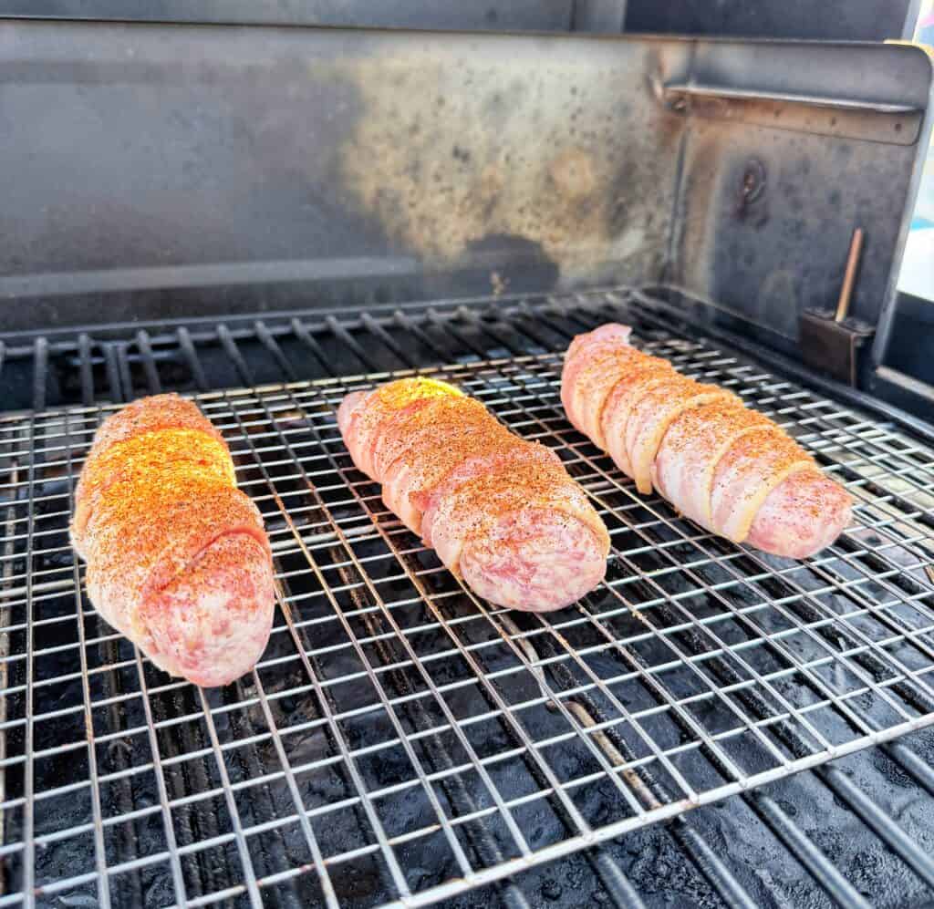 Three seasoned bacon-wrapped bratwursts cooking on a grill.