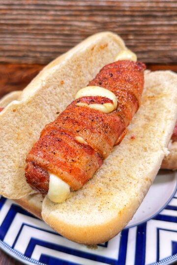 Bacon-wrapped sausage stuffed with cheese in a sliced bun on a patterned plate.