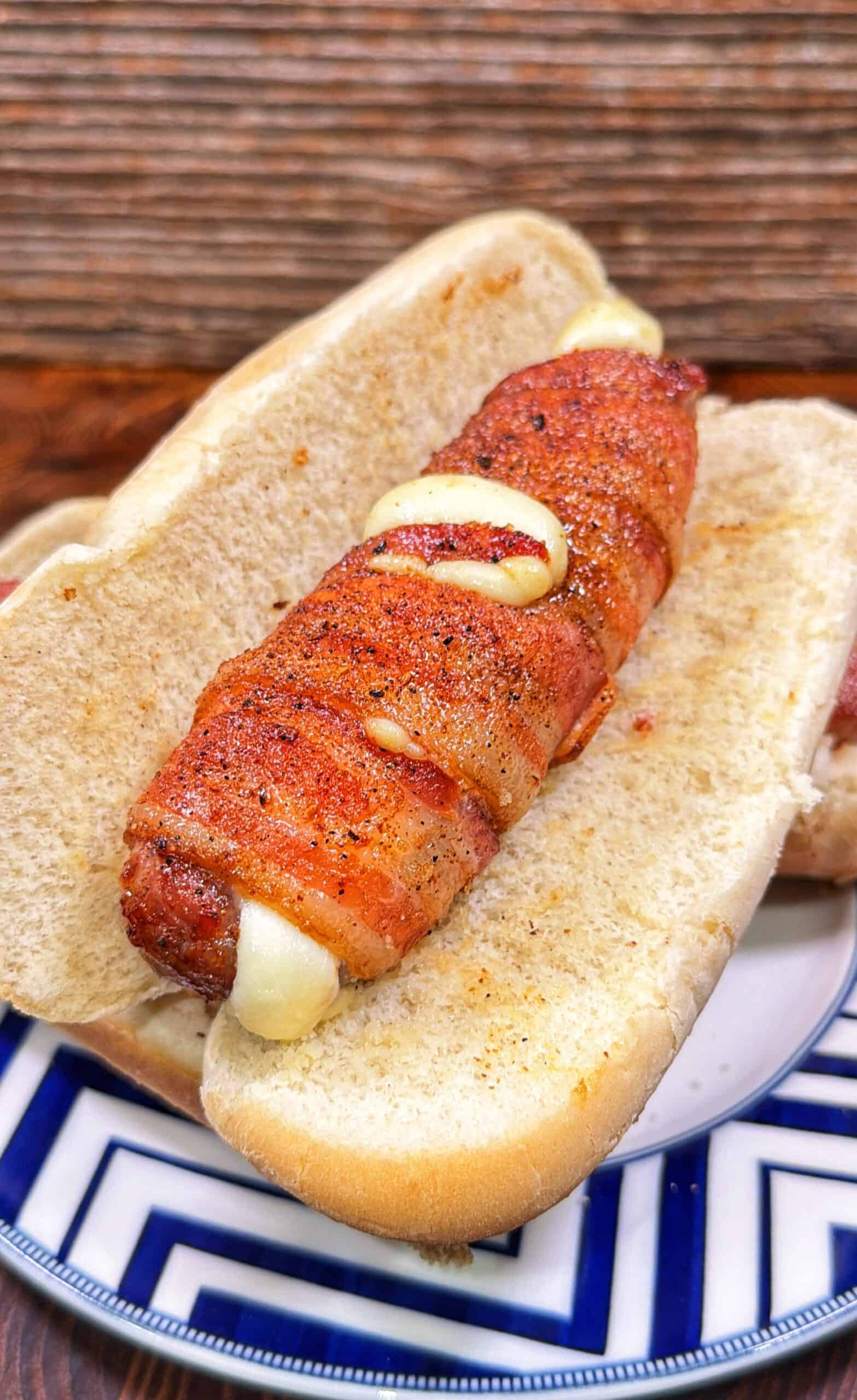 Bacon-wrapped sausage stuffed with cheese in a sliced bun on a patterned plate.