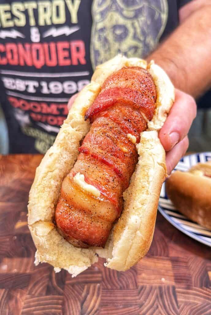 A hand holds a bun filled with a bacon-wrapped bratwurst.