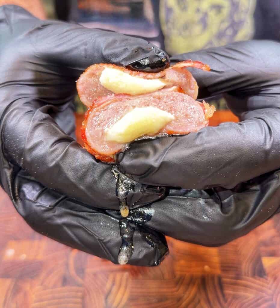 Gloved hands hold a sliced bacon-wrapped bratwurst stuffed with melted cheese.