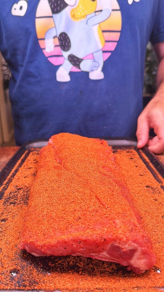 A seasoned rack of ribs covered in red spice rub on a baking tray.