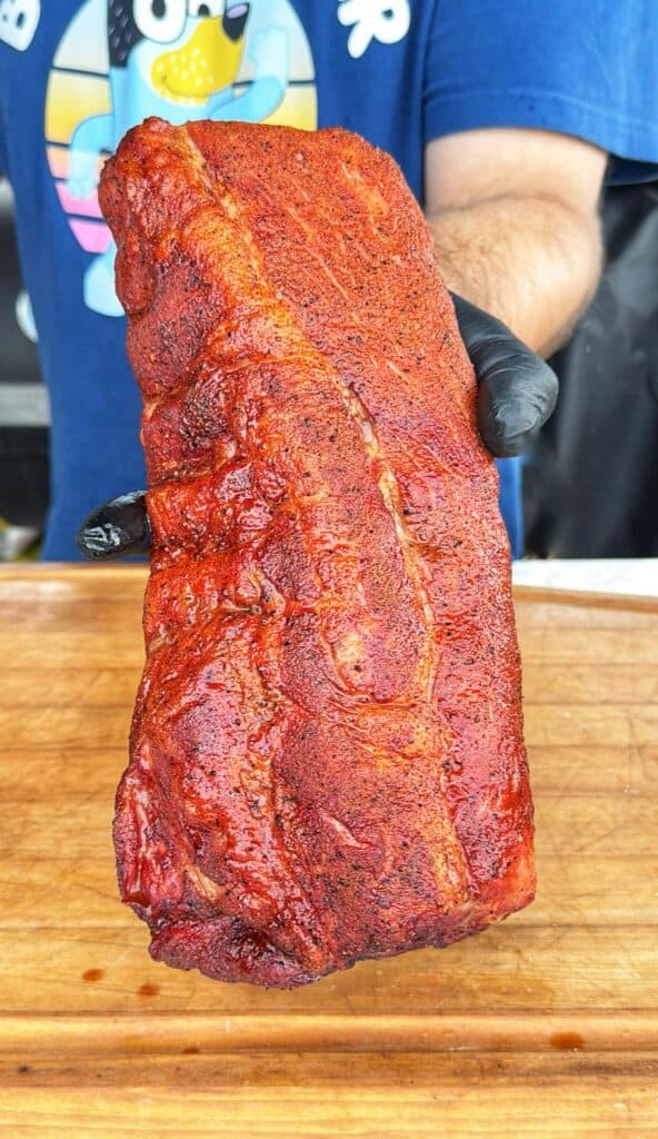 A person wearing black gloves holds a seasoned rack of smoked ribs upright over a wooden cutting board.