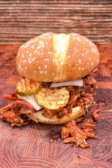 Pulled pork rib sandwich topped with pickles and onions on a wooden surface, with shredded pork spilling onto the board.