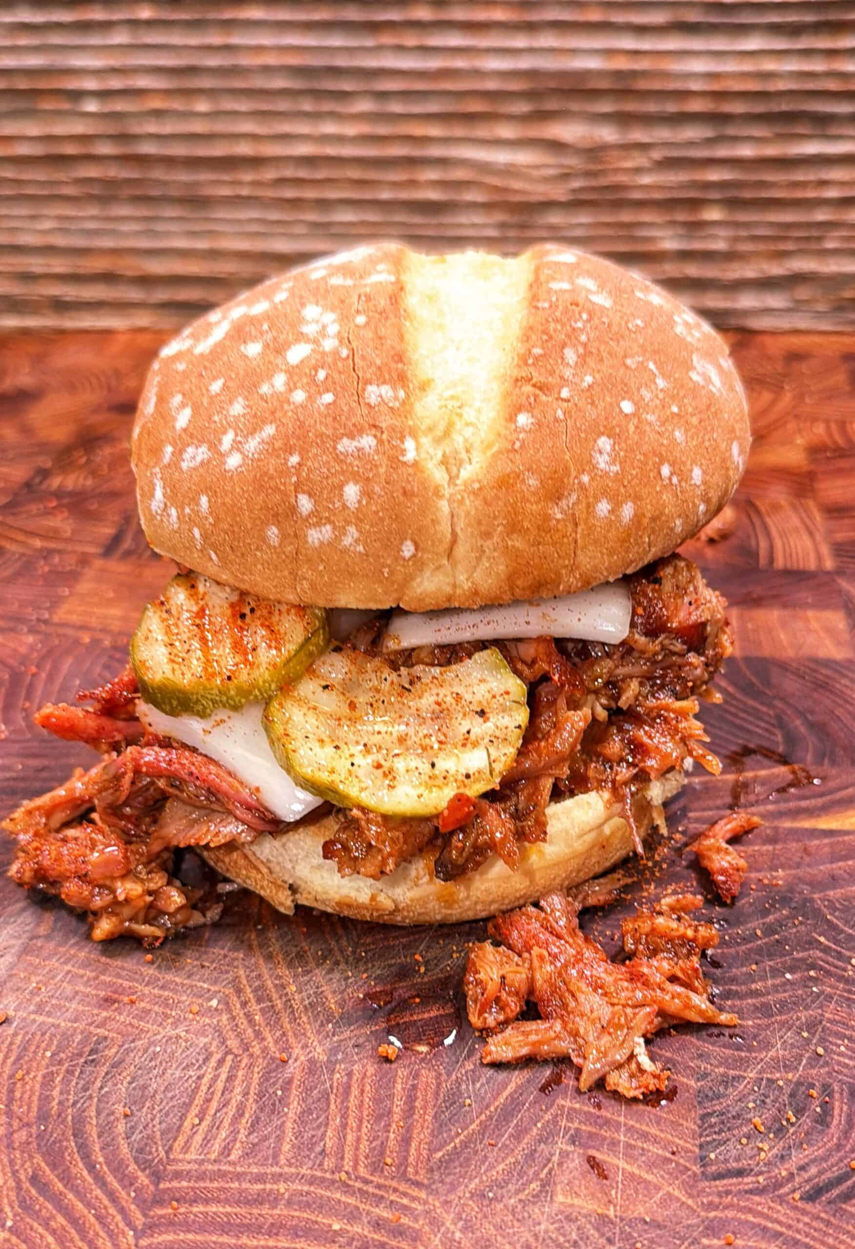 Pulled pork rib sandwich topped with pickles and onions on a wooden surface, with shredded pork spilling onto the board.