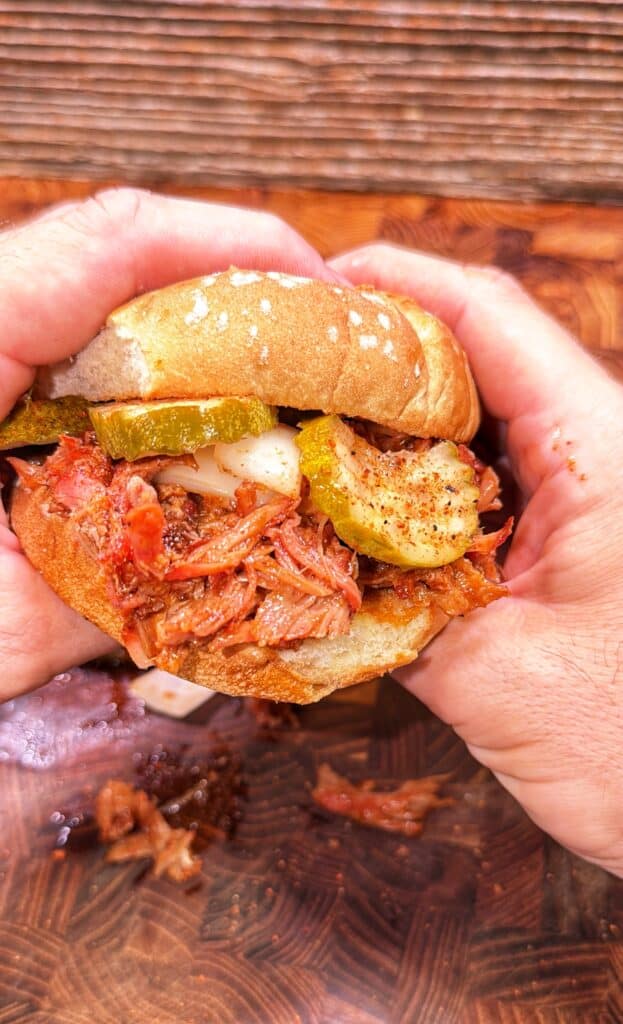 A person holds a pulled pork rib sandwich topped with pickles and onions.
