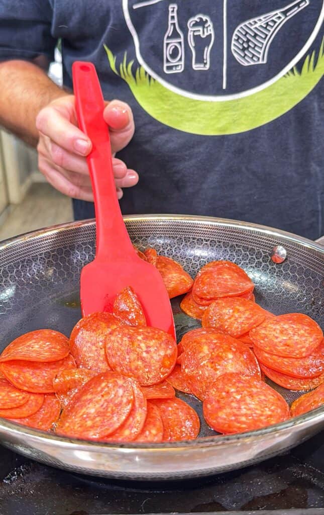 Sliced pepperoni cooking in a frying pan as it is stirred with a spatula.