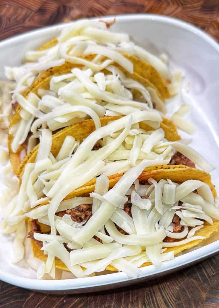 Three hard-shell pizza tacos filled with pizza meat and topped with shredded mozzarella in a baking dish.