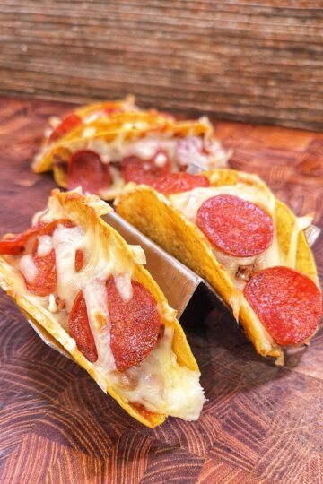 Three pizza tacos filled with melted mozzarella and pepperoni slices in a metal taco holder.