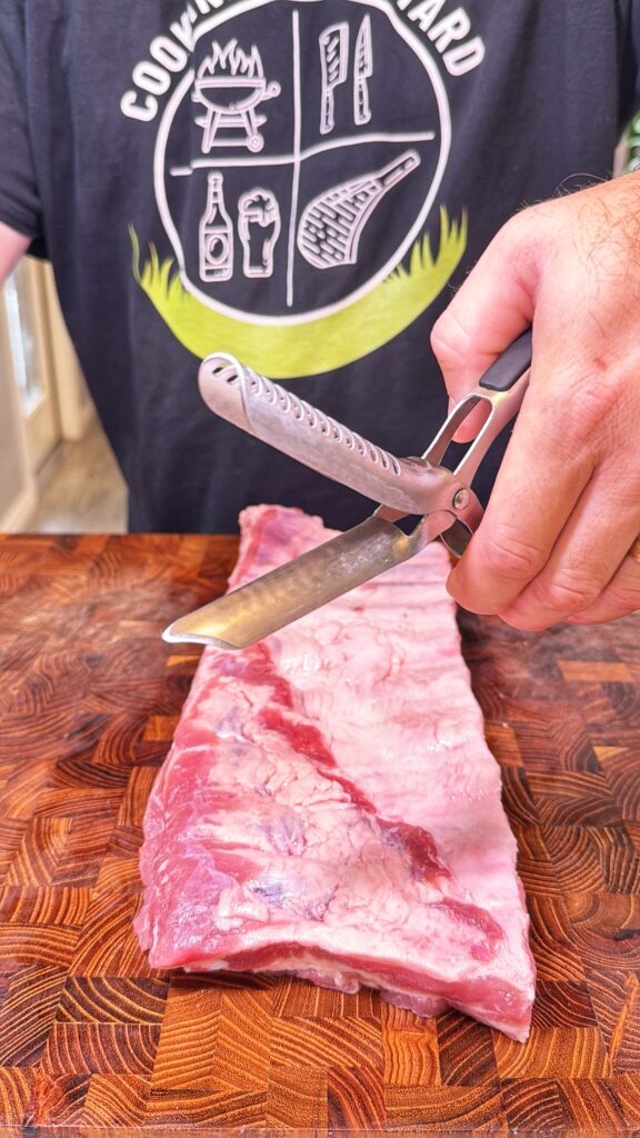A person trims a rack of raw pork ribs with kitchen shears on a wooden cutting board.