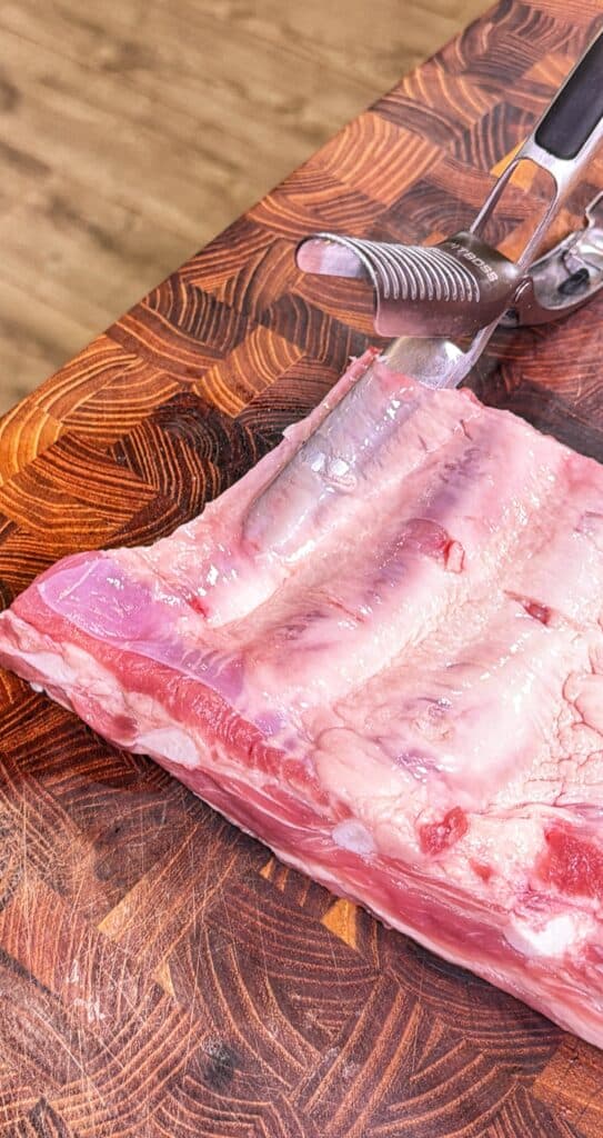 A rack of raw pork ribs on a wooden cutting board with a meat tenderizer resting on top.