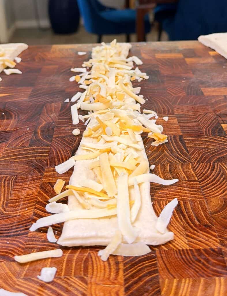 Shredded cheese spread in a line on a strip of raw dough on a wooden cutting board.