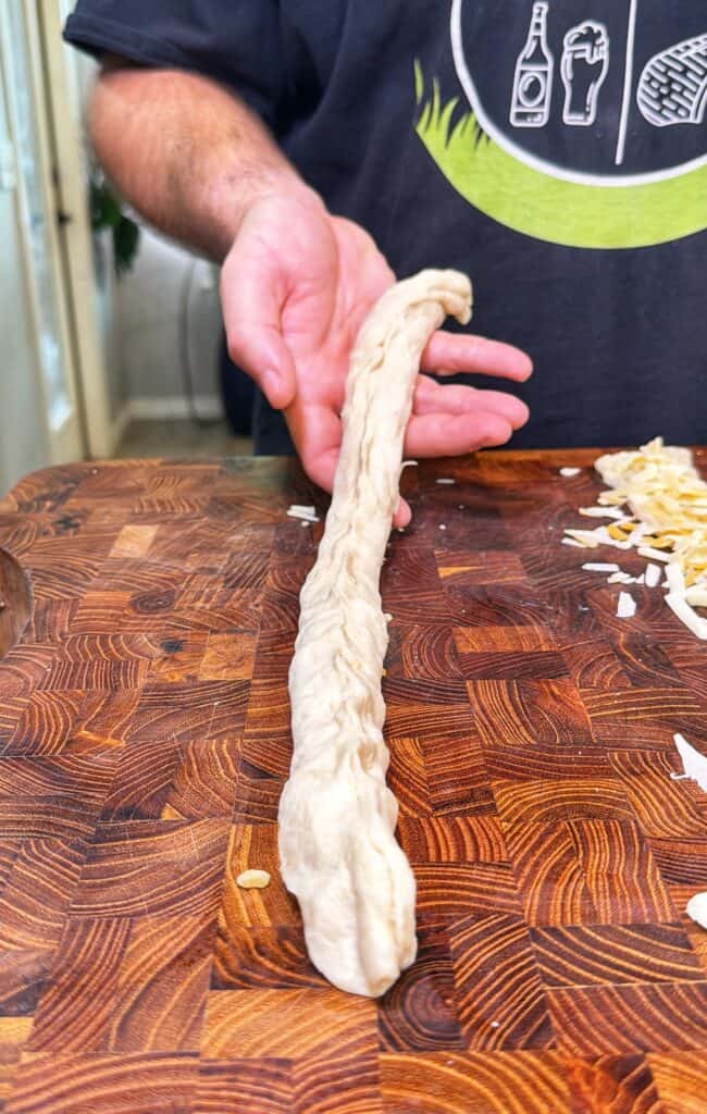 A hand holds a long twisted piece of raw dough on a wooden cutting board, with shredded cheese nearby.