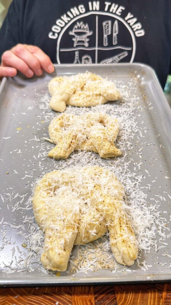 Three unbaked pretzel-shaped dough pieces topped with shredded cheese on a baking sheet.