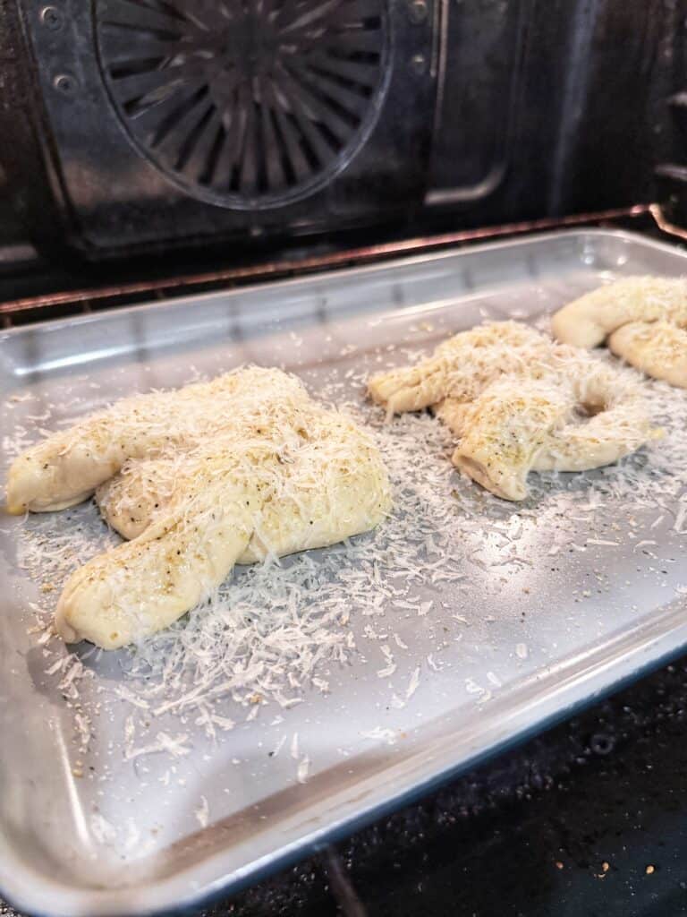 Unbaked pretzel-shaped dough sprinkled with grated cheese and herbs on a baking sheet inside an oven.