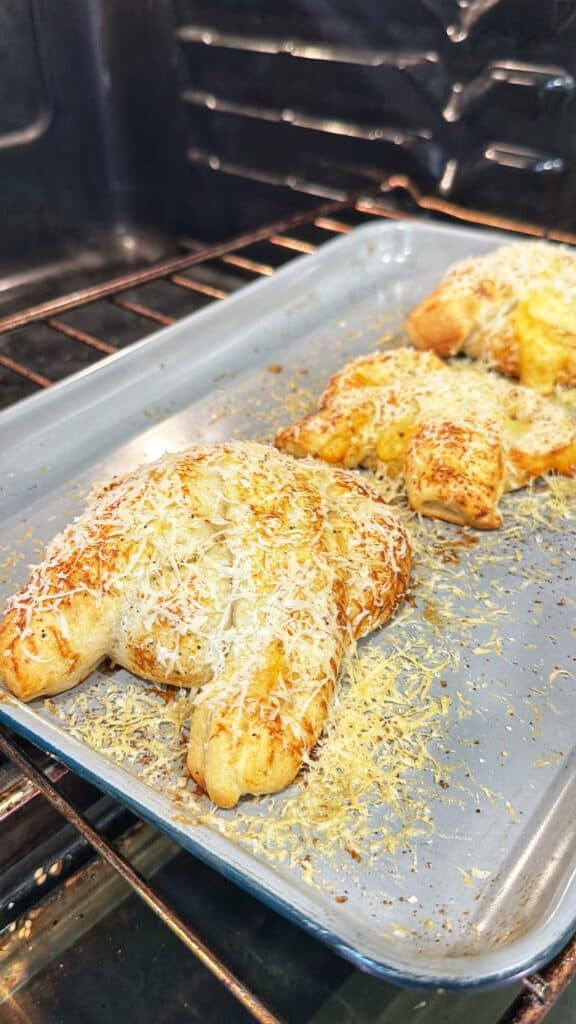 Two cheese-topped pretzels baking on a metal tray inside an oven.