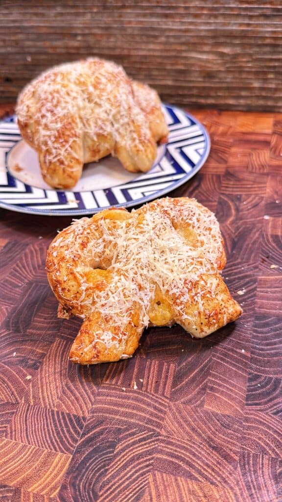 A baked cheese-topped pretzel on a wooden board, with another pretzel on a plate in the background.