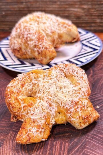 A close-up of a baked pretzel topped with shredded cheese on a wooden surface, with another cheese-topped pretzel on a plate in the background.