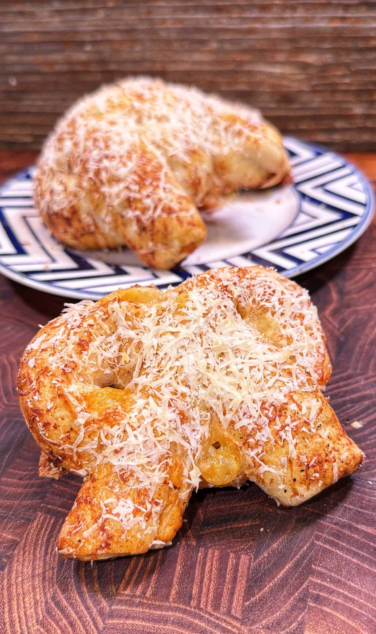 A close-up of a baked pretzel topped with shredded cheese on a wooden surface, with another cheese-topped pretzel on a plate in the background.